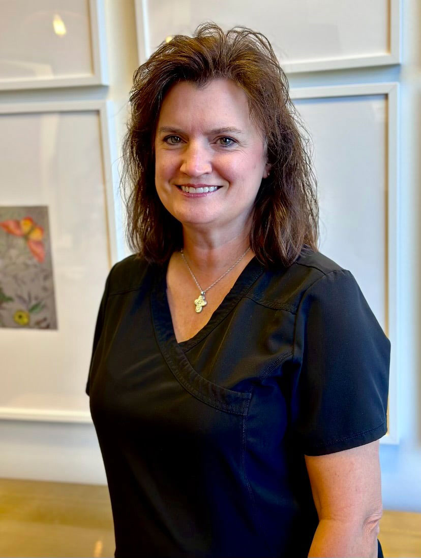 A woman wearing a black shirt and white pants stands in front of framed pictures on a wall.